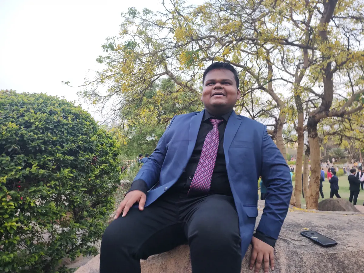Aditya smiling, sitting on a rock under trees