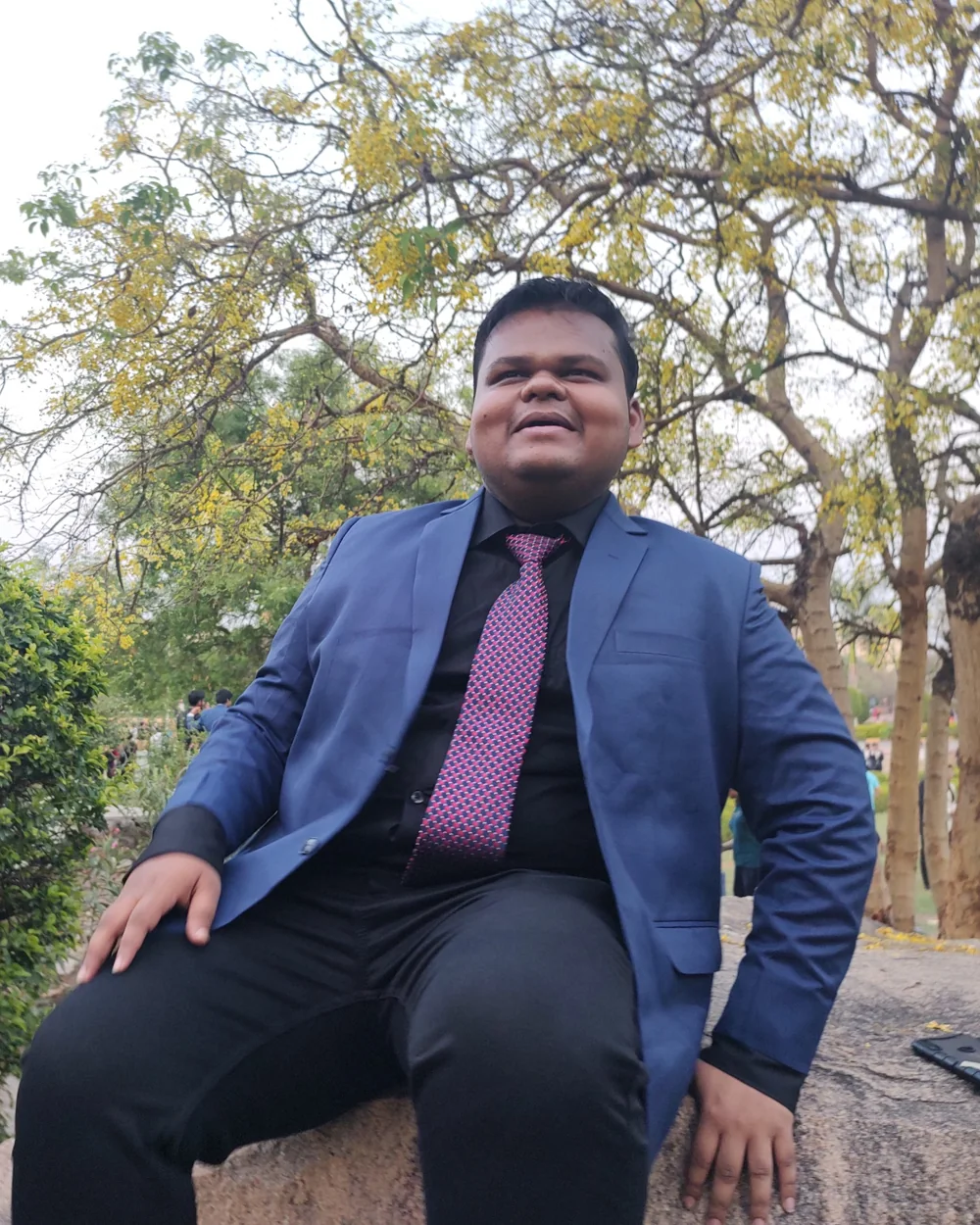 Aditya in a blue suit, laughing, sitting on a rock under a flowering tree